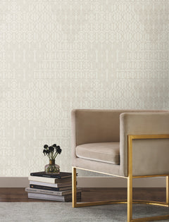 A sleek beige armchair with a gold frame sits beside a stack of books and a small vase on a rug, set against a wall adorned with the Modern Chandelier Wallpaper by York Wallcoverings.