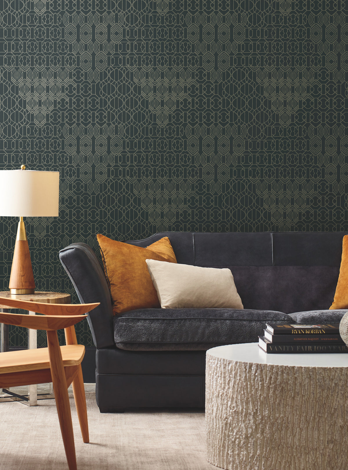 Modern living room featuring York Wallcoverings’ Modern Chandelier Wallpaper, a black sofa accented with tan and beige pillows, a round white textured coffee table with books stacked on top, a wooden side table, and a gold table lamp.