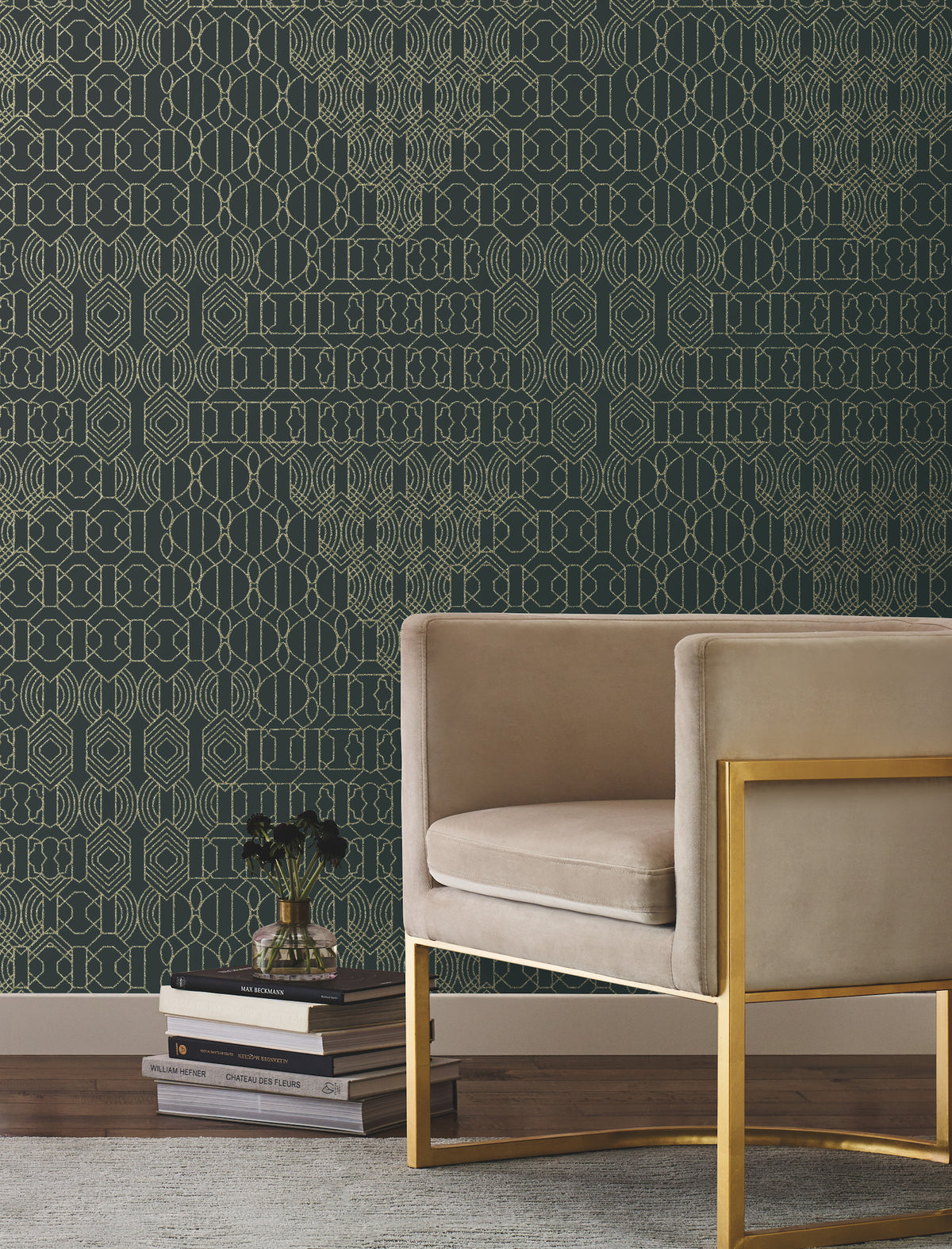 The Modern Chandelier Wallpaper by York Wallcoverings in geometric gold and dark green sets a striking backdrop behind a modern beige velvet armchair with a gold frame, positioned next to a stack of books and a small black vase with flowers, all placed atop a light textured rug on a wooden floor.