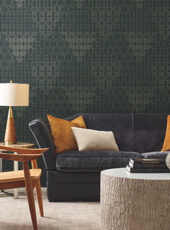 Modern living room with a dark gray sofa, tan and cream pillows, a round textured coffee table, stacked books, a wooden side table, and a gold lamp beside an accent wall decorated with the Modern Chandelier Wallpaper by York Wallcoverings.