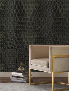A modern beige velvet chair with a gold metal frame sits on a gray rug next to stacked books. The background features the York Wallcoverings Modern Chandelier Wallpaper, showcasing an intricate gold geometric pattern on a black wall.