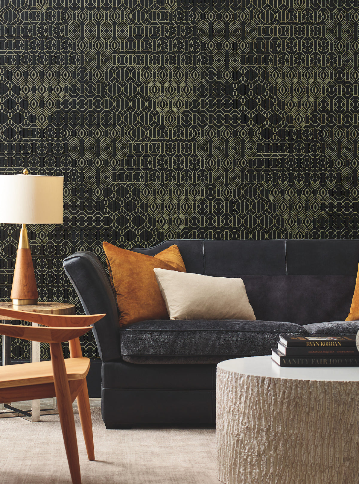 A contemporary living room featuring a dark sofa, orange and cream pillows, a wooden side table, lamp, and a round textured coffee table. The wall is adorned with the Modern Chandelier Wallpaper by York Wallcoverings, showcasing a striking black and gold geometric pattern.