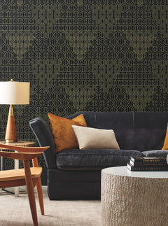 A contemporary living room featuring a dark sofa, orange and cream pillows, a wooden side table, lamp, and a round textured coffee table. The wall is adorned with the Modern Chandelier Wallpaper by York Wallcoverings, showcasing a striking black and gold geometric pattern.