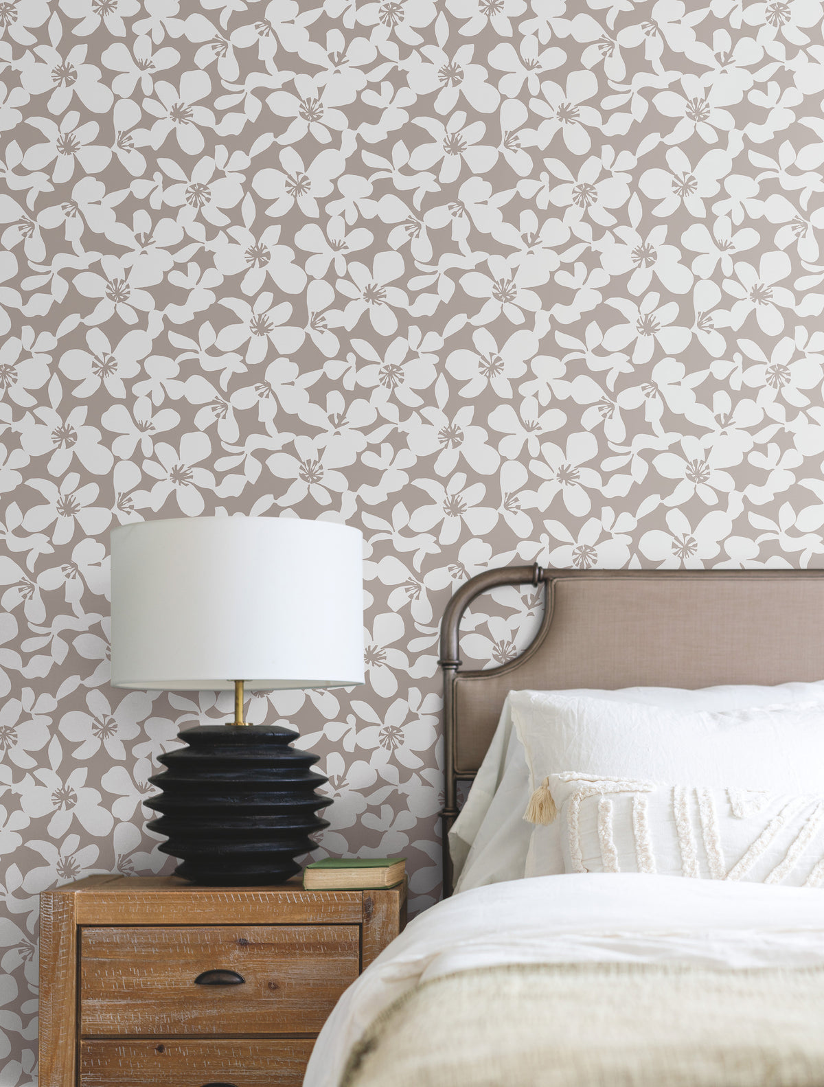 A bedside table with a black lamp sits next to a bed with white bedding and a beige headboard, set against a wall adorned with the Free Spirit Wallpaper by York Wallcoverings.