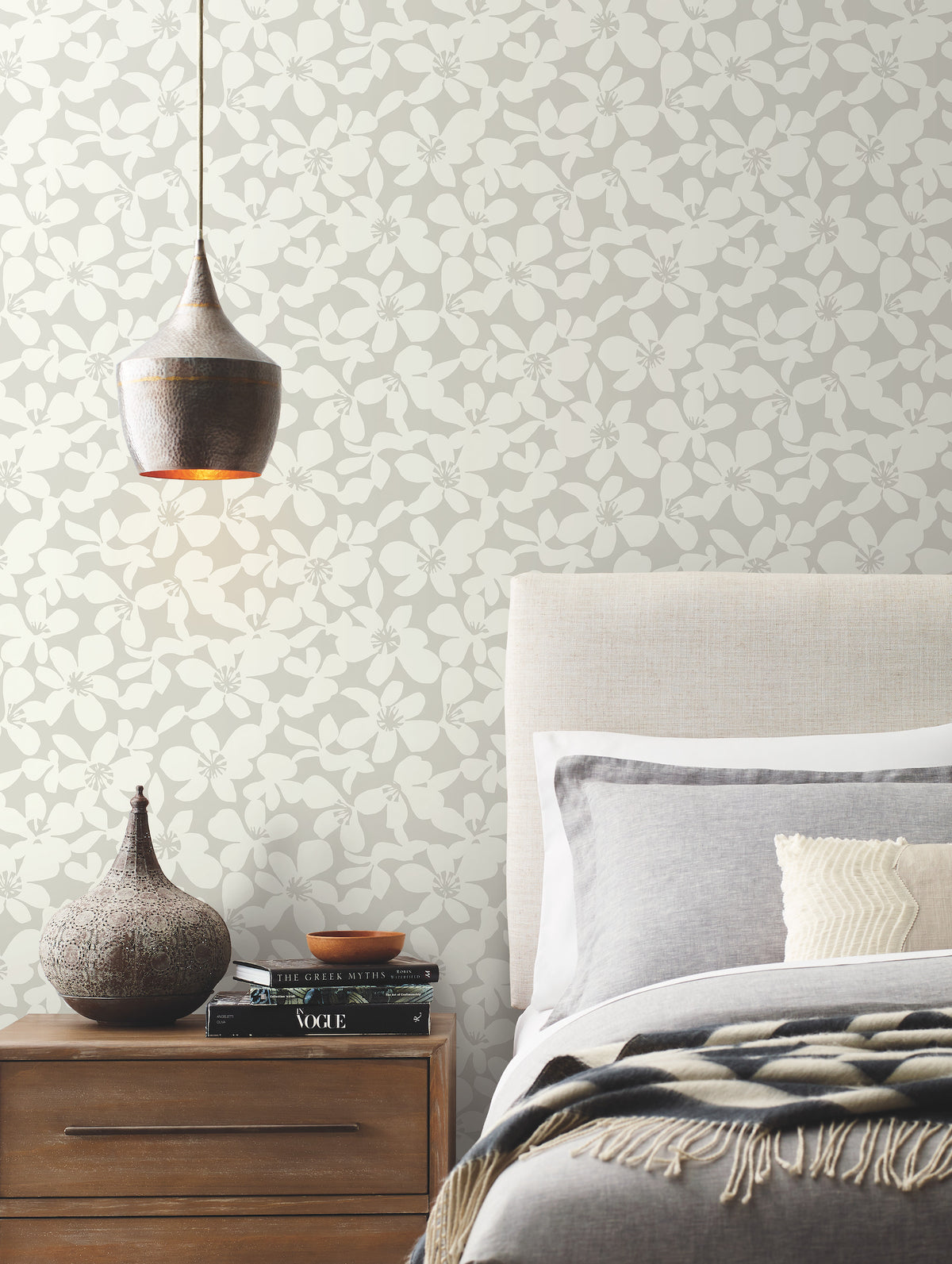 A modern bedroom featuring York Wallcoverings’ Free Spirit Wallpaper in gray floral, a beige upholstered headboard, gray bedding, a wooden nightstand adorned with books and vases, and a copper pendant light hanging above.
