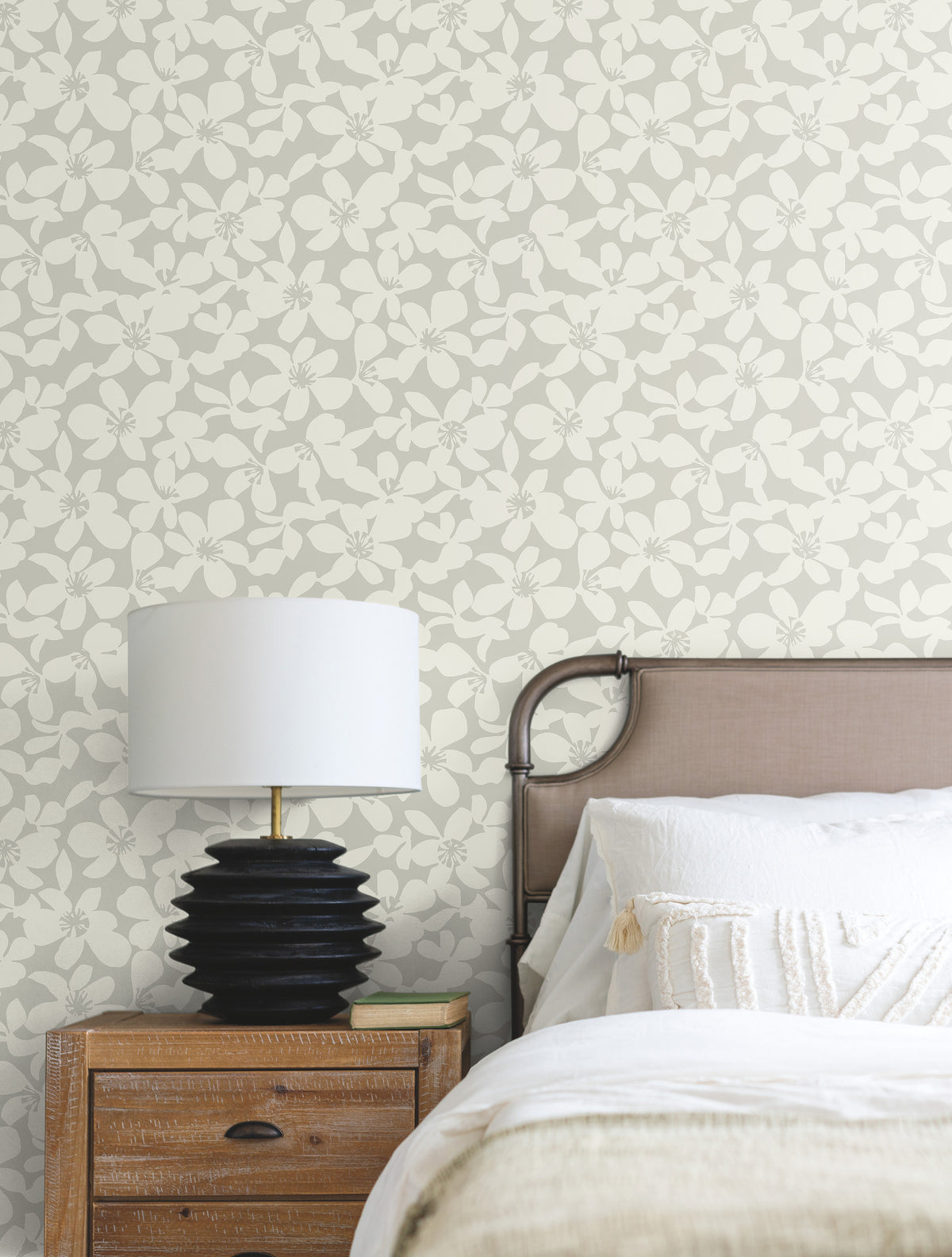 A bedside table with a black lamp sits next to a bed with white bedding and a beige headboard, set against a wall covered in York Wallcoverings’ Free Spirit Wallpaper.