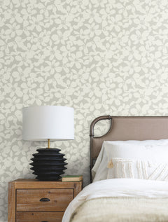 A bedside table with a black lamp sits next to a bed with white bedding and a beige headboard, set against a wall covered in York Wallcoverings’ Free Spirit Wallpaper.