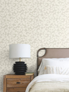A nightstand with a black lamp and a book beside a bed with a beige tufted headboard, white bedding, and York Wallcoverings’ Free Spirit Wallpaper in the background.