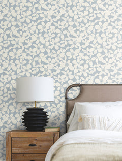 A nightstand with a black lamp and a white shade sits beside a bed with white bedding, set against a wall adorned with York Wallcoverings’ Free Spirit Wallpaper in a blue and cream butterfly pattern.