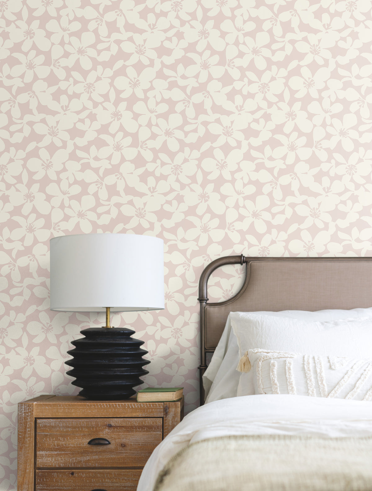 A bedside table with a black lamp and book sits next to a bed with white bedding, while the wall features York Wallcoverings’ Free Spirit Wallpaper in light pink and cream floral patterns.