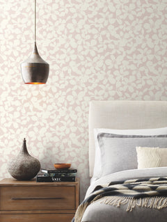 A cozy bedroom corner featuring York Wallcoverings’ Free Spirit Wallpaper in soft neutral tones, complemented by a gray bed, textured pillows, a wooden nightstand, a decorative vase, and a hanging metallic lamp.