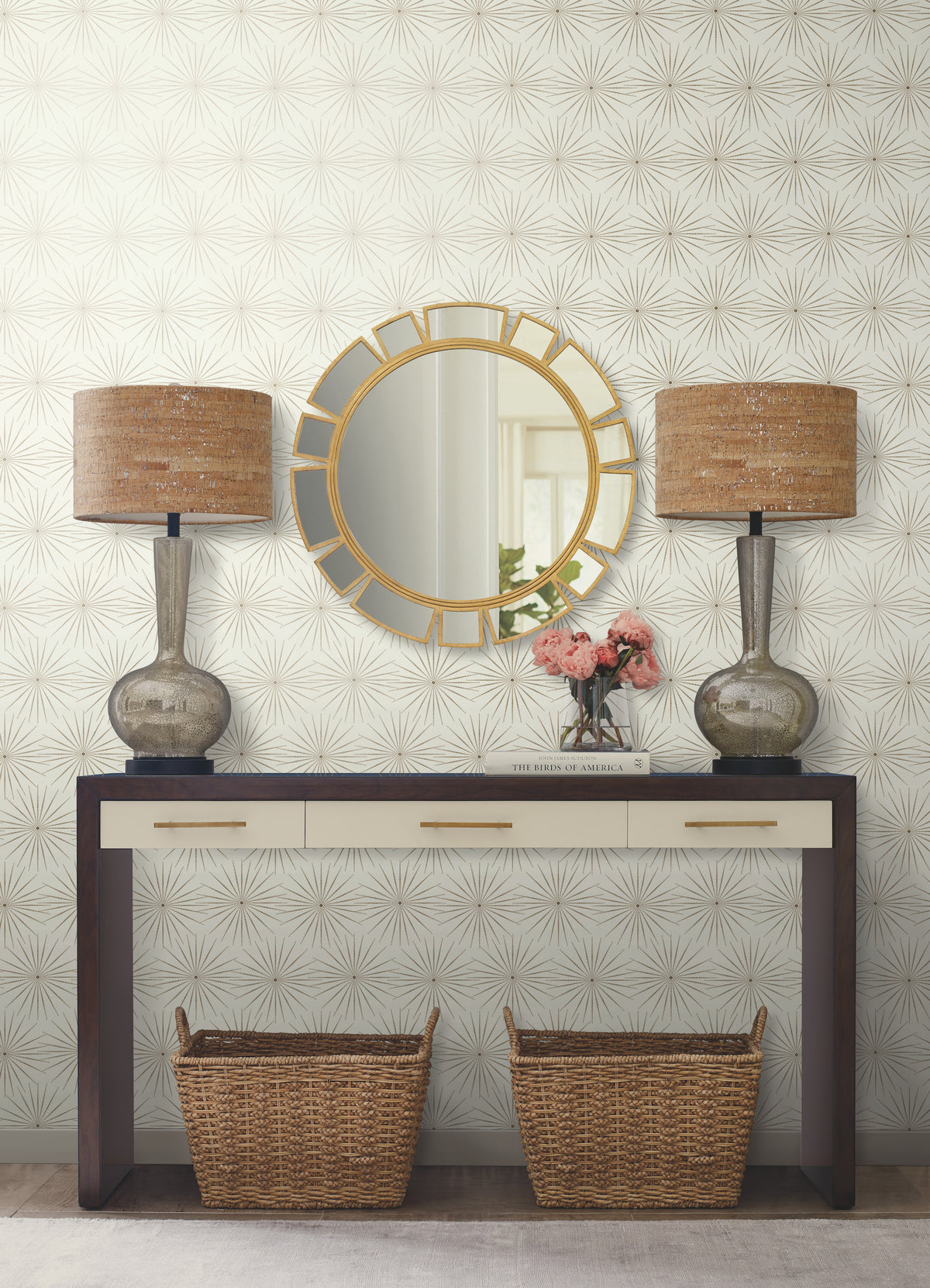 A console table with two wicker baskets beneath, two lamps, a vase of pink flowers, and a round gold mirror above, is set against York Wallcoverings’ Starlight Wallpaper.