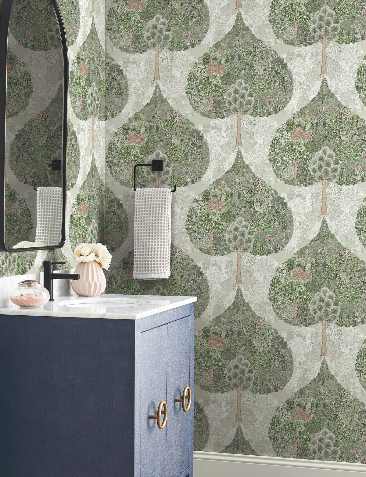 A bathroom featuring York Wallcoverings’ Mystic Forest Wallpaper adorned with green trees and small animals, complemented by a blue vanity, marble countertop, oval mirror, black towel holder with a towel, and a pink vase filled with flowers.