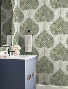 A bathroom featuring York Wallcoverings’ Mystic Forest Wallpaper adorned with green trees and small animals, complemented by a blue vanity, marble countertop, oval mirror, black towel holder with a towel, and a pink vase filled with flowers.