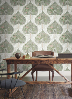 A wooden desk with a brown leather chair sits on a woven rug in front of a wall covered with Mystic Forest Wallpaper by York Wallcoverings, featuring lush green leafy patterns. An open book and decorative objects are displayed atop the desk.