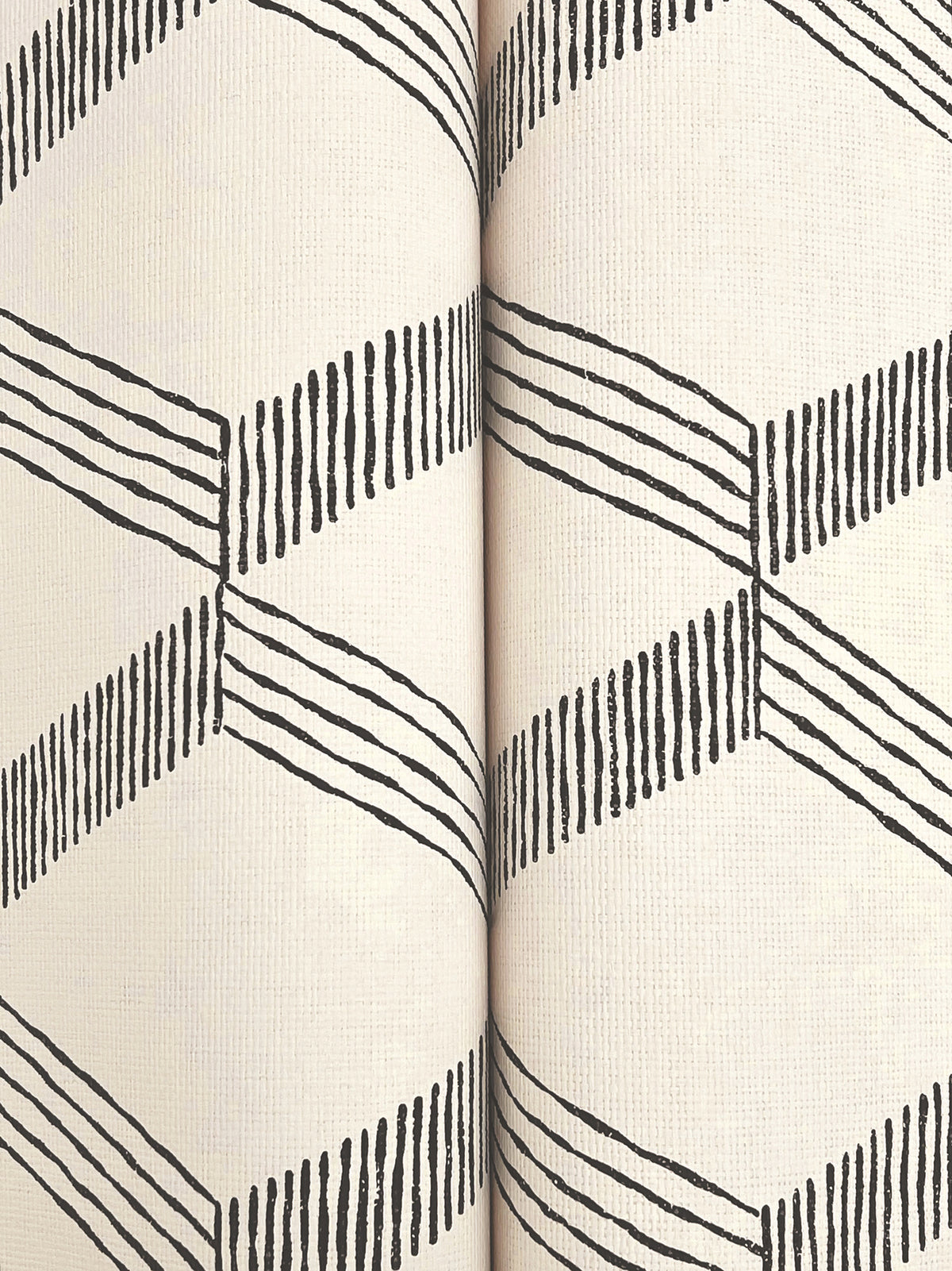 Close-up of two rolls of Palisades Paperweave Wallpaper by York Wallcoverings, featuring a cream background and a repeating black geometric zigzag pattern made from angled lines.