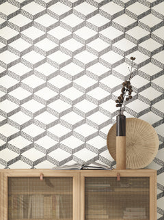 Palisades Paperweave Wallpaper by York Wallcoverings, featuring a black and white cube pattern, serves as a stylish backdrop behind a light wooden cabinet. Atop the cabinet are a round fan, a tall vase with dried branches, and a book titled MAP.