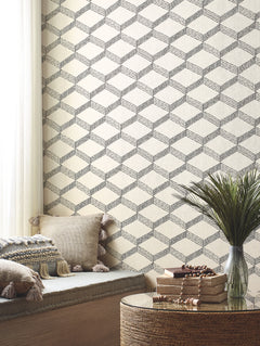 A cozy nook features beige cushions with tassels, a round wicker table topped with books and beads, and Palisades Paperweave Wallpaper by York Wallcoverings in a geometric pattern. Soft light filters through white curtains.