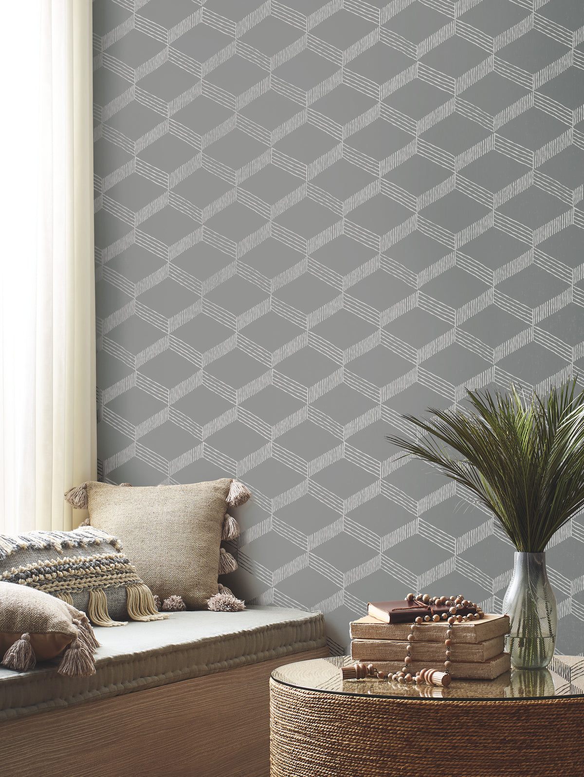 A cozy corner featuring York Wallcoverings’ Palisades Paperweave Wallpaper in a gray geometric pattern, a cushioned bench adorned with tasseled pillows, a round table stacked with books, and a vase of green foliage placed near a window dressed in sheer curtains.