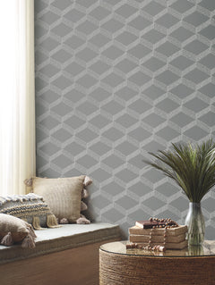 A cozy corner featuring York Wallcoverings’ Palisades Paperweave Wallpaper in a gray geometric pattern, a cushioned bench adorned with tasseled pillows, a round table stacked with books, and a vase of green foliage placed near a window dressed in sheer curtains.