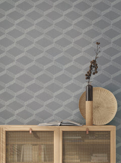 Behind a wooden cabinet with mesh doors, an open book, a round wooden object, and a tall vase with dried branches on top, the Palisades Paperweave Wallpaper by York Wallcoverings in gray geometric pattern adds visual interest to the space.