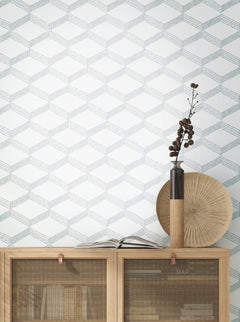 A light wooden cabinet adorned with books and decor sits against a white wall covered in Palisades Paperweave Wallpaper by York Wallcoverings, which features a subtle geometric cube pattern. On top of the cabinet are a round fan-like object, a tall vase with dried branches, and an open book.