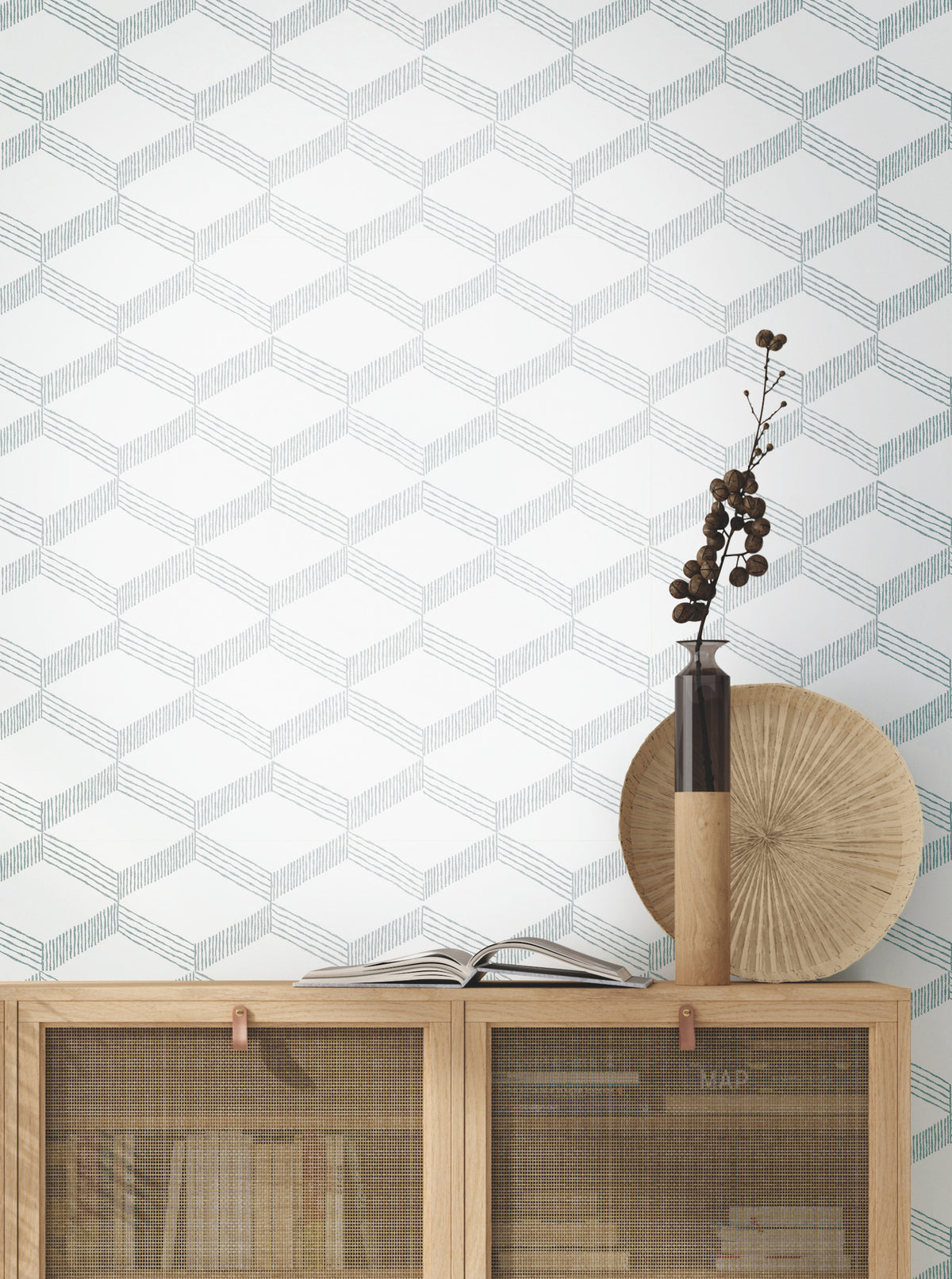 A wooden cabinet with books and decor sits against a wall covered in York Wallcoverings’ Palisades Paperweave Wallpaper, featuring a geometric pattern. On top are a tall vase with branches, a round textured plate, and an open book.