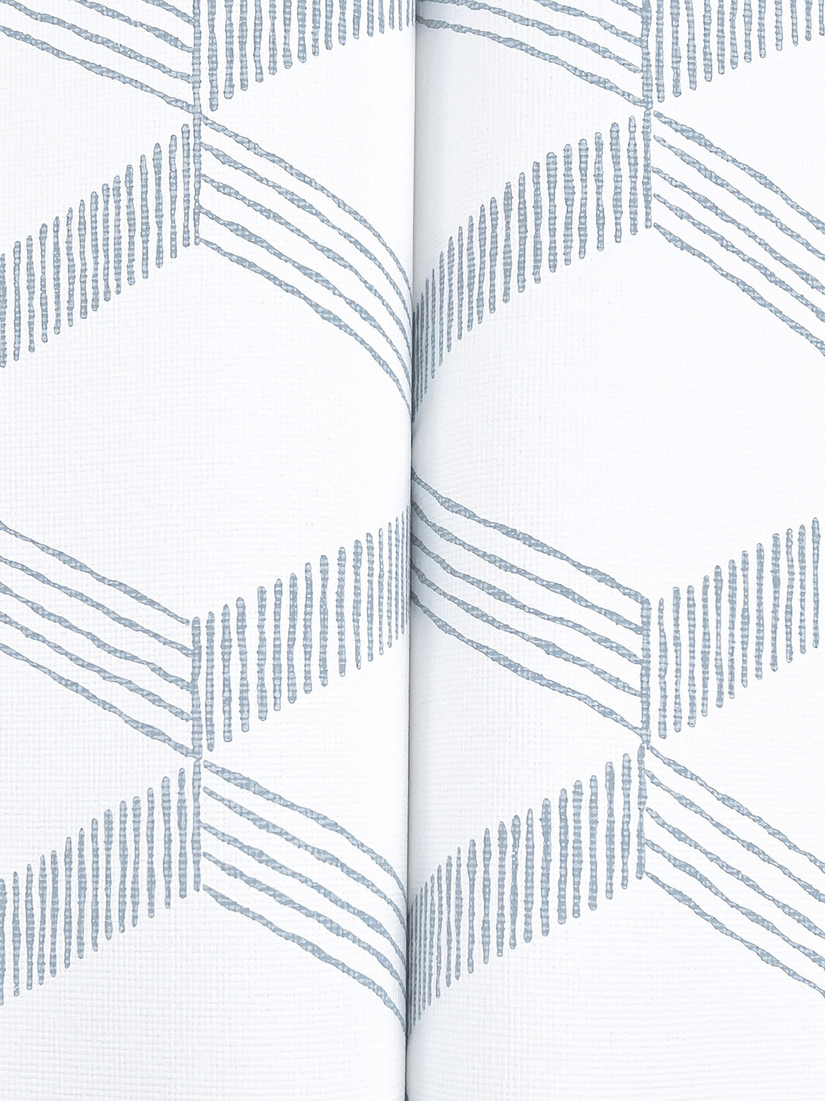 Close-up of Palisades Paperweave Wallpaper by York Wallcoverings in light gray, featuring a repeating geometric pattern of diagonal lines that create zigzags. A vertical seam is visible down the center where two pieces meet, set against a white background.