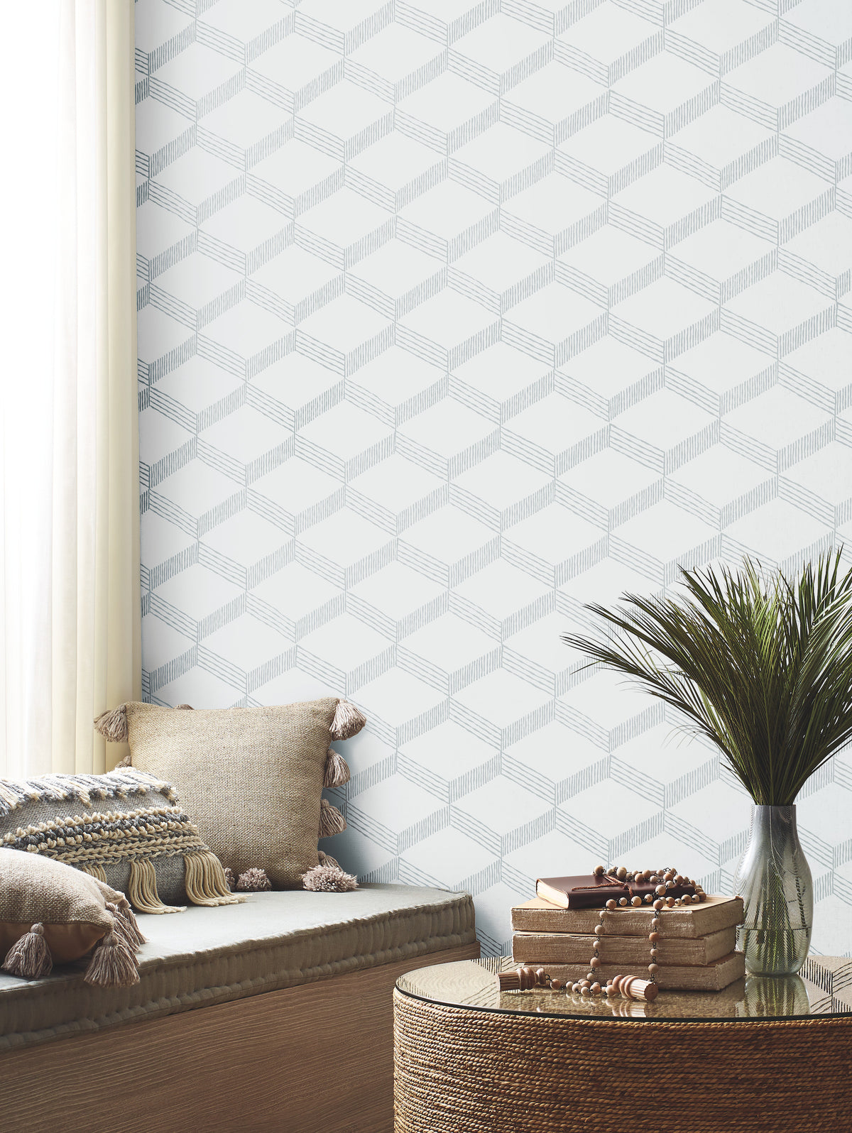 A cozy reading nook with neutral cushions, a round woven table holding books and beads, and a green plant is set against the soft gray and white geometric pattern of York Wallcoverings’ Palisades Paperweave Wallpaper. Light pours in through a sheer curtain.