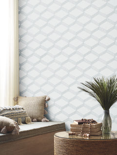 A cozy corner featuring textured pillows, a cushioned bench, a round wicker table adorned with books and beads, a vase filled with tall green grass, and Palisades Paperweave Wallpaper by York Wallcoverings in soft tones. Gentle light filters through a white curtain.