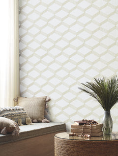 A cozy nook featuring beige tasseled pillows, a small potted plant, and stacked books on a round woven table sits by a window with sheer white curtains, while the wall is adorned with York Wallcoverings’ Palisades Paperweave Wallpaper.