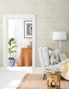 A cozy living room featuring York Wallcoverings’ Laurel Damask Wallpaper, a white armchair with throw pillows, a gold side table, a lamp, and a woven rug. Through the doorway, a fiddle-leaf fig in a blue pot and a wicker console are visible.