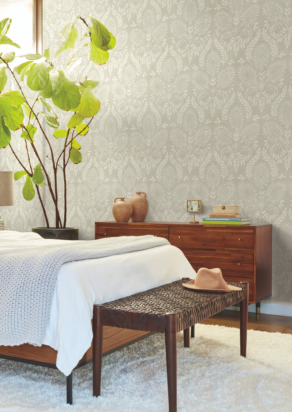 A cozy bedroom features a wooden bed, woven bench, and large leafy plant. The Laurel Damask Wallpaper by York Wallcoverings adds textured elegance to the gray wall, while a dresser with vases, a hat, and neutral decor completes the calm, stylish atmosphere.