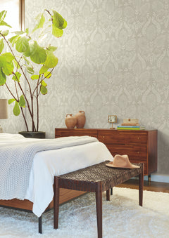 A cozy bedroom features a wooden bed, woven bench, and large leafy plant. The Laurel Damask Wallpaper by York Wallcoverings adds textured elegance to the gray wall, while a dresser with vases, a hat, and neutral decor completes the calm, stylish atmosphere.
