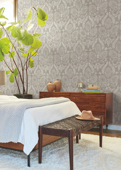 Mid-century modern bedroom featuring a wooden bed, a woven bench topped with a beige hat, and York Wallcoverings’ Laurel Damask Wallpaper in gray. A dresser with decor, a large leafy plant, and soft natural light enhance the cozy space.