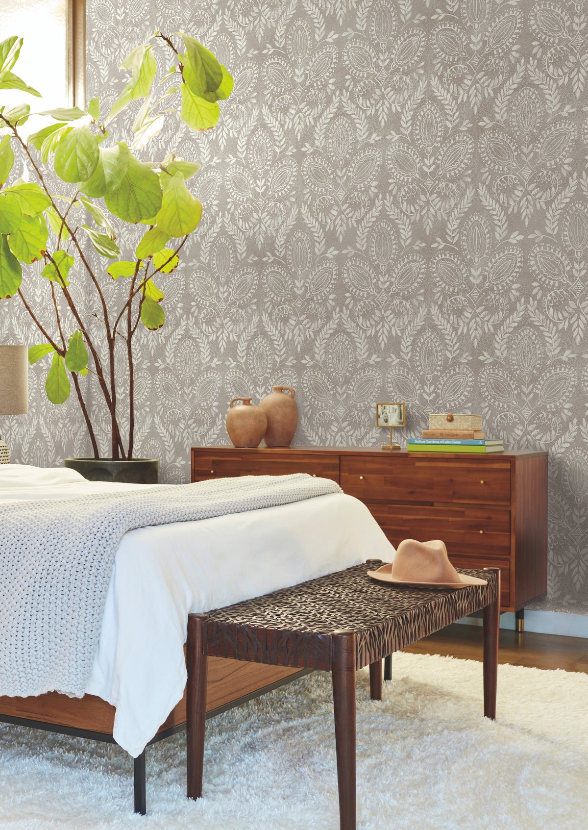 A cozy bedroom adorned with Laurel Damask Wallpaper by York Wallcoverings, featuring a leafy plant, wooden furniture, a bed topped with a knit blanket, and a bench displaying a hat. Soft natural light fills the space.