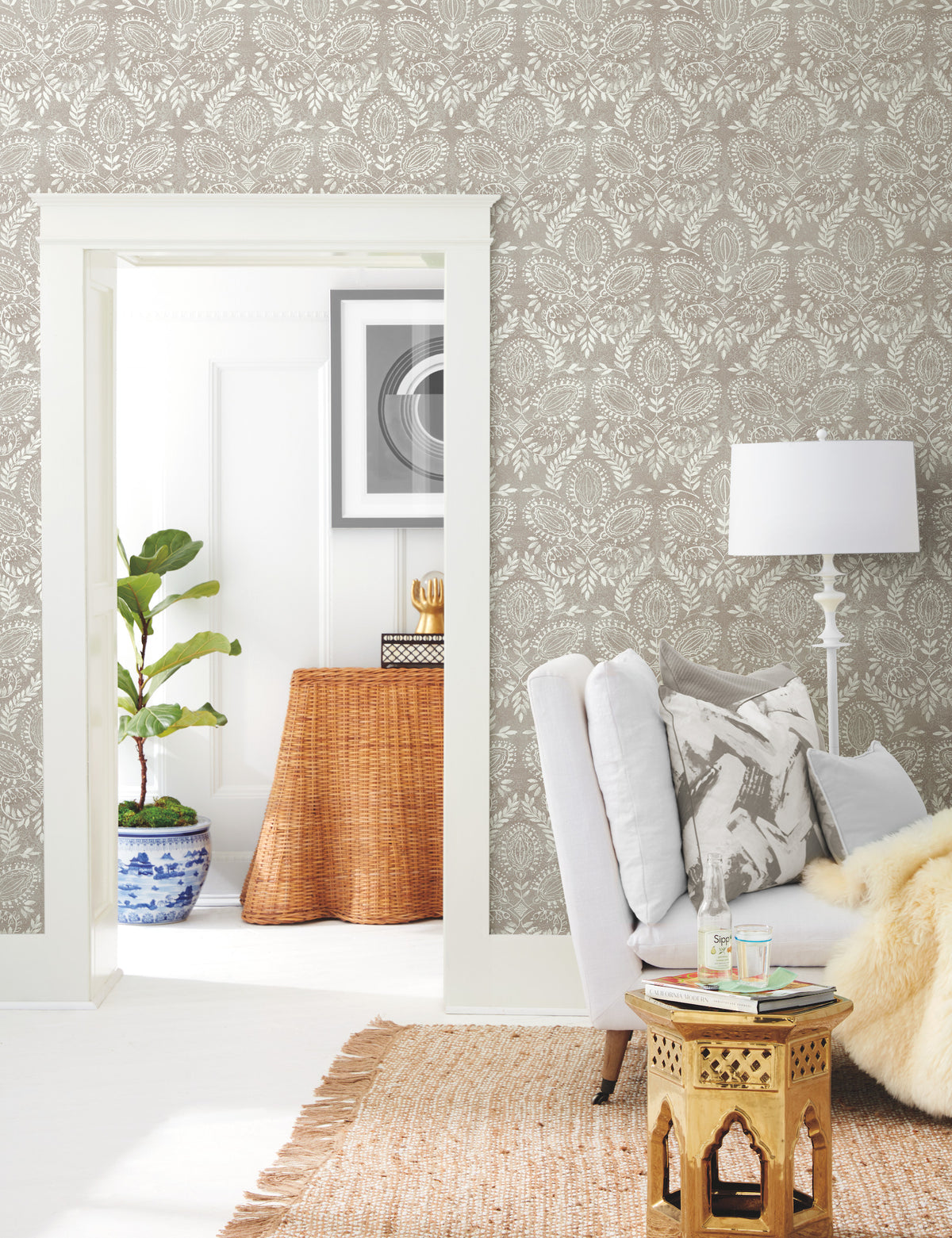 A cozy living room with a white armchair, small gold side table, floor lamp, and leafy plant. Laurel Damask Wallpaper from York Wallcoverings adds pattern to the walls, while a woven rug completes the decor; a wicker console and framed art are visible in the next room.