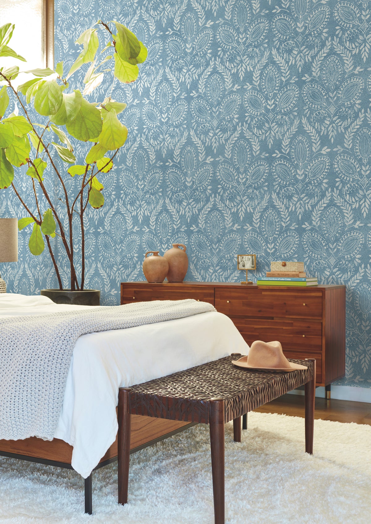 A cozy bedroom featuring York Wallcoverings’ Laurel Damask Wallpaper in blue, a wooden bed with a white blanket, a brown woven bench topped with a hat, a leafy green plant, a wooden dresser, and decor items arranged on a white rug.