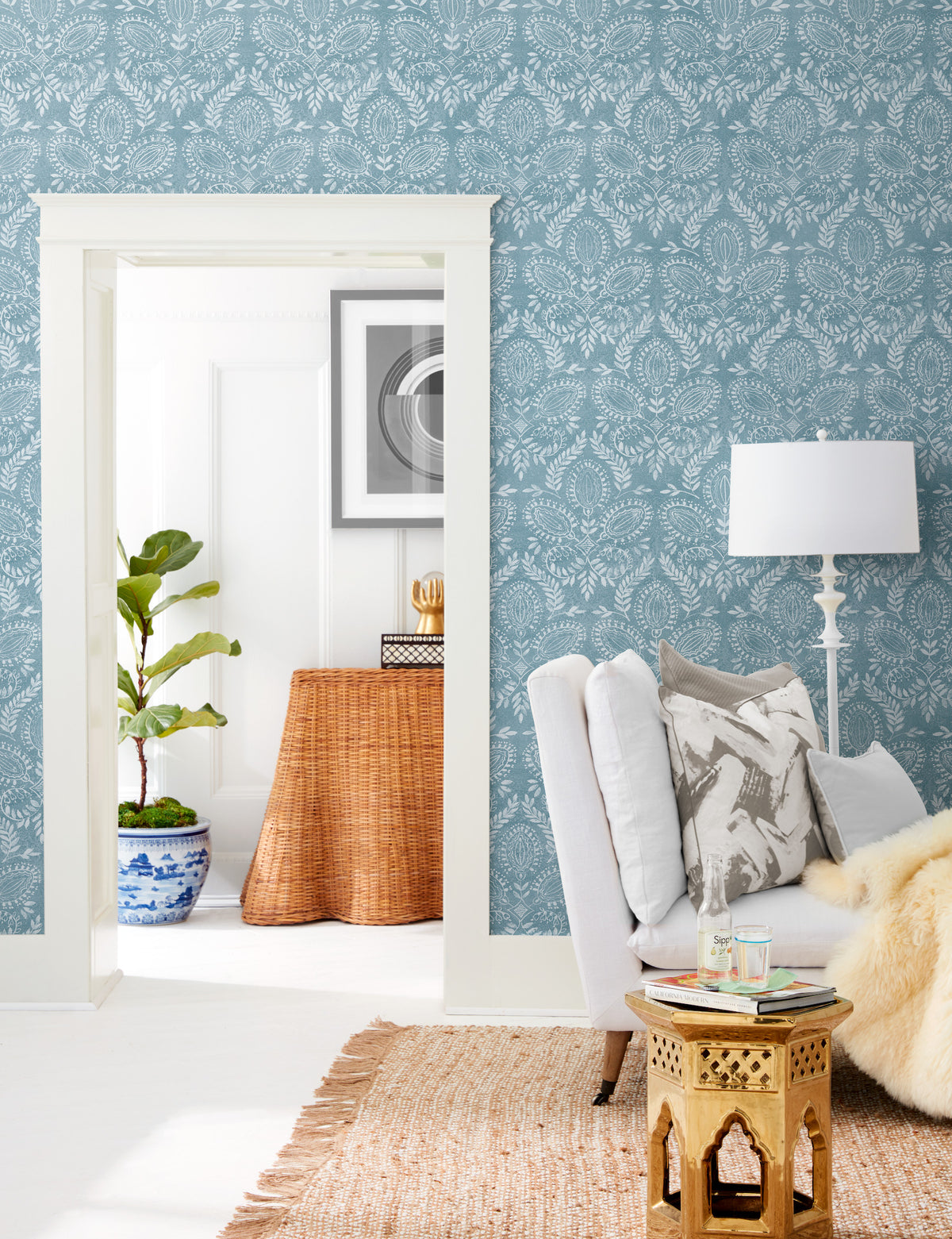 Bright living room featuring York Wallcoverings’ Laurel Damask Wallpaper in blue, a white armchair, a gold side table, a floor lamp, a jute rug, a potted plant, and a wicker console visible through the doorway.