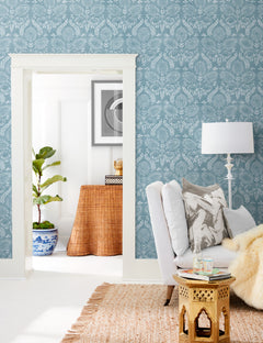 Bright living room featuring York Wallcoverings’ Laurel Damask Wallpaper in blue, a white armchair, a gold side table, a floor lamp, a jute rug, a potted plant, and a wicker console visible through the doorway.