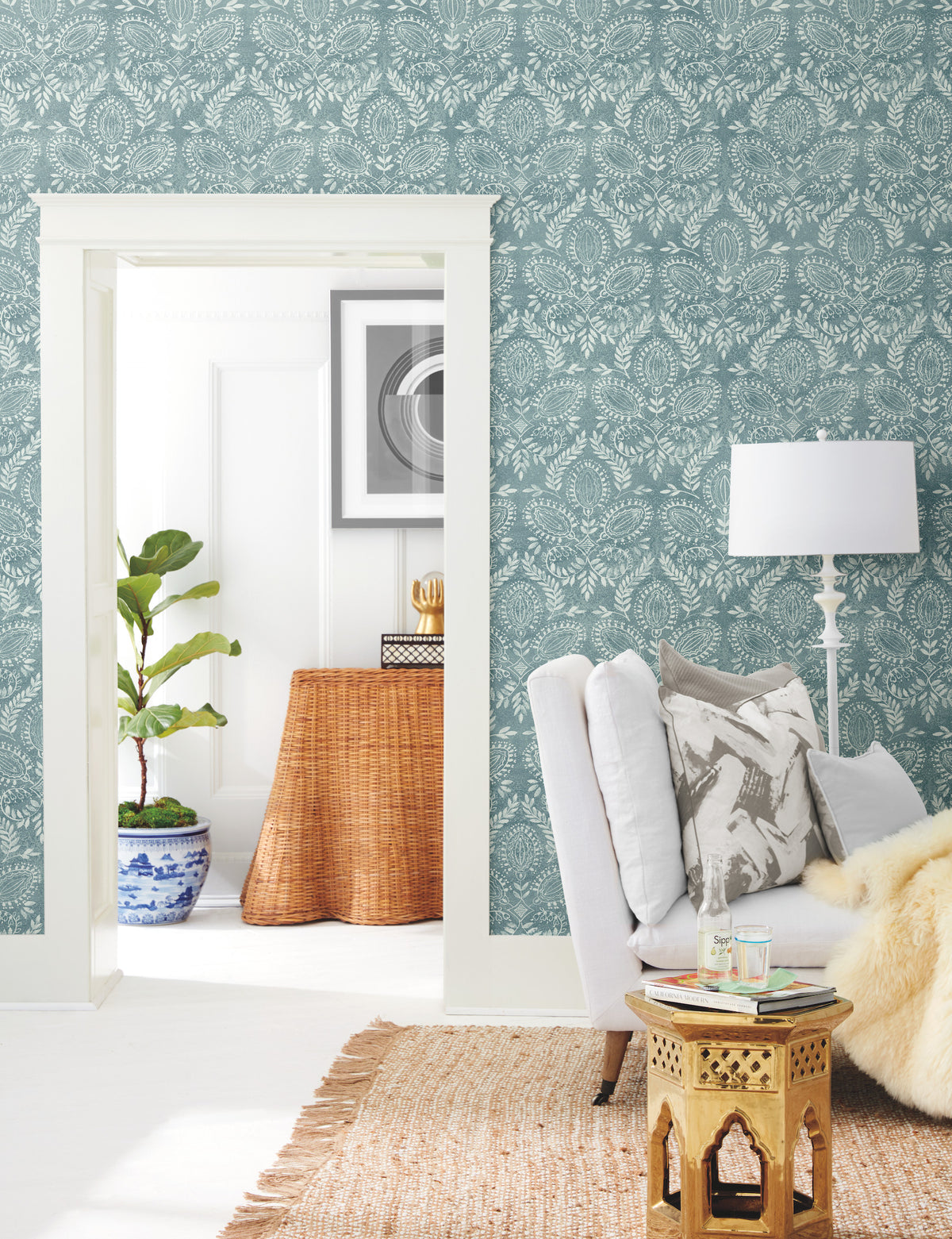 A cozy living room featuring Laurel Damask Wallpaper by York Wallcoverings, a white armchair, a gold side table, and a woven rug. Through the doorway, there’s a wicker console table, a plant in a blue pot, and framed art on the wall.