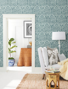 A cozy living room featuring Laurel Damask Wallpaper by York Wallcoverings, a white armchair, a gold side table, and a woven rug. Through the doorway, there’s a wicker console table, a plant in a blue pot, and framed art on the wall.