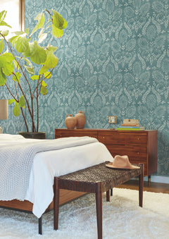Mid-century modern bedroom featuring York Wallcoverings’ Laurel Damask Wallpaper in blue, a wooden bed and dresser, a woven bench topped with a hat, a white rug, potted plant, and natural light streaming in from the left.