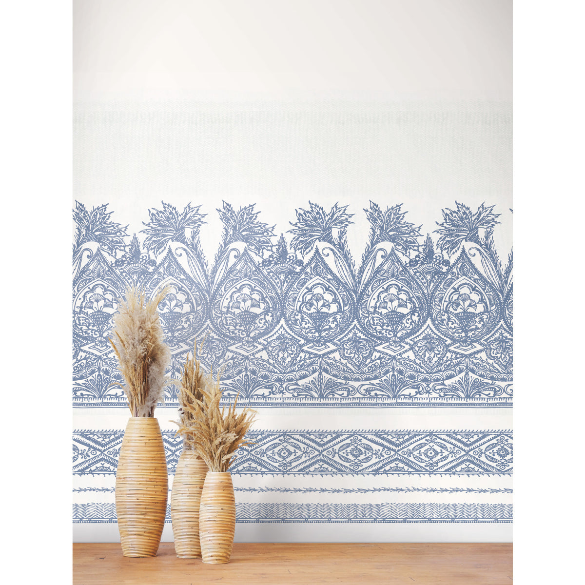 Three woven vases filled with dried grasses sit on a wooden surface in front of a wall adorned with the Henna Wall Mural by York Wallcoverings, featuring intricate blue and white floral and geometric patterns.