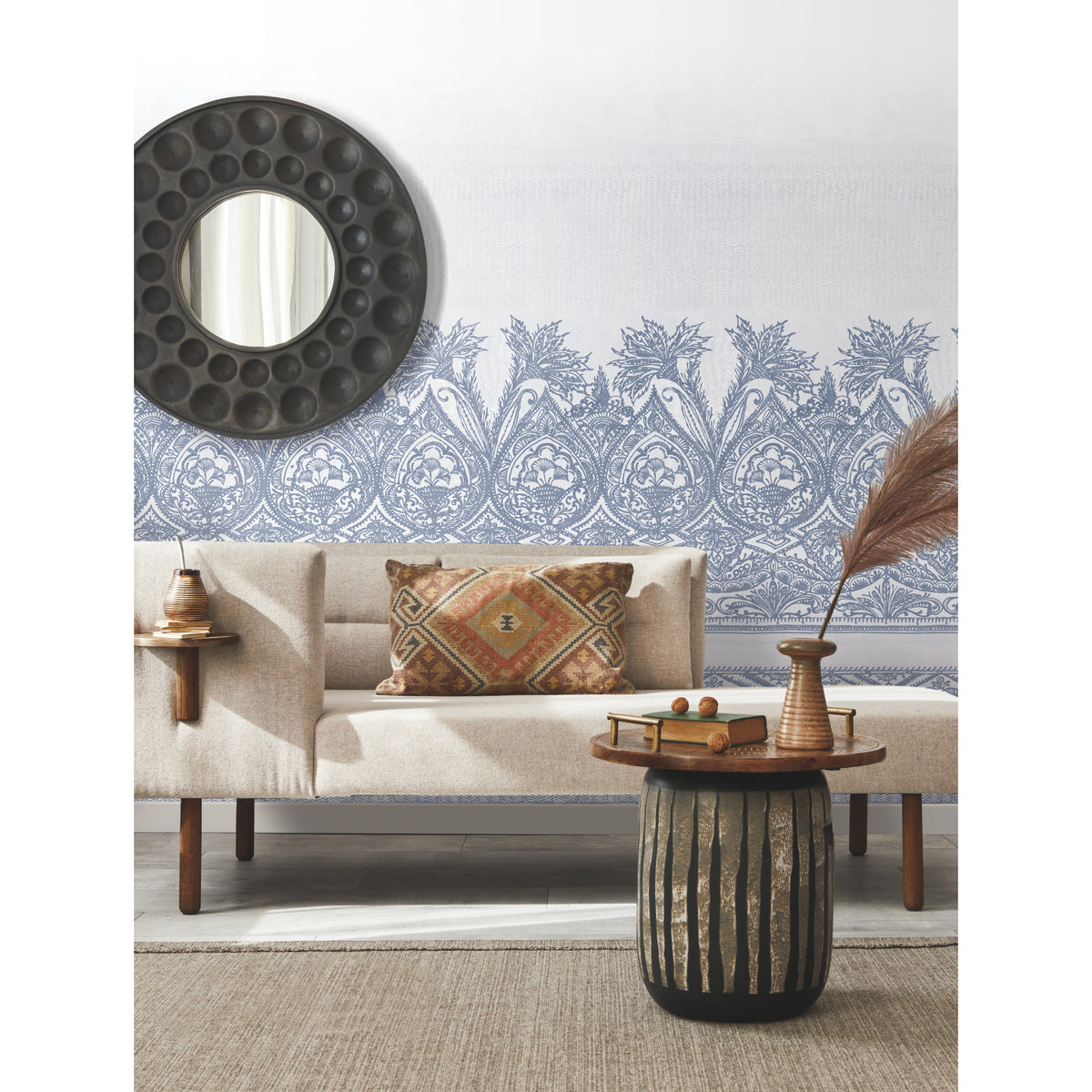 A modern living room featuring a beige sofa, a round mirror on the wall, York Wallcoverings’ Henna Wall Mural as patterned blue wallpaper, a rustic side table with books and vases, and a textured rug on the floor.