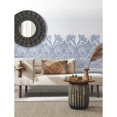 A modern living room featuring a beige sofa, a round mirror on the wall, York Wallcoverings’ Henna Wall Mural as patterned blue wallpaper, a rustic side table with books and vases, and a textured rug on the floor.