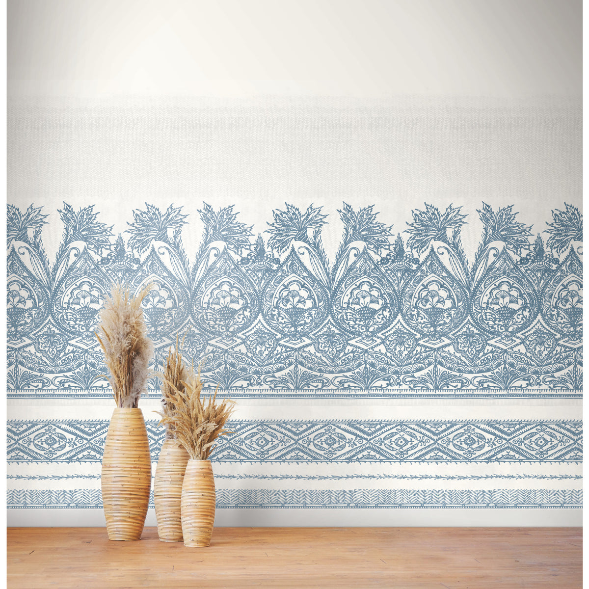 Two tall wicker vases filled with dried pampas grass stand on a wooden floor in front of a white wall adorned with the Henna Wall Mural by York Wallcoverings, featuring intricate blue floral patterns.