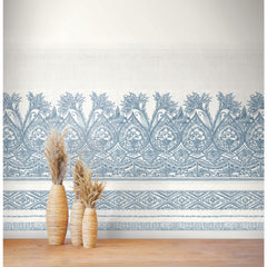 Two tall wicker vases filled with dried pampas grass stand on a wooden floor in front of a white wall adorned with the Henna Wall Mural by York Wallcoverings, featuring intricate blue floral patterns.