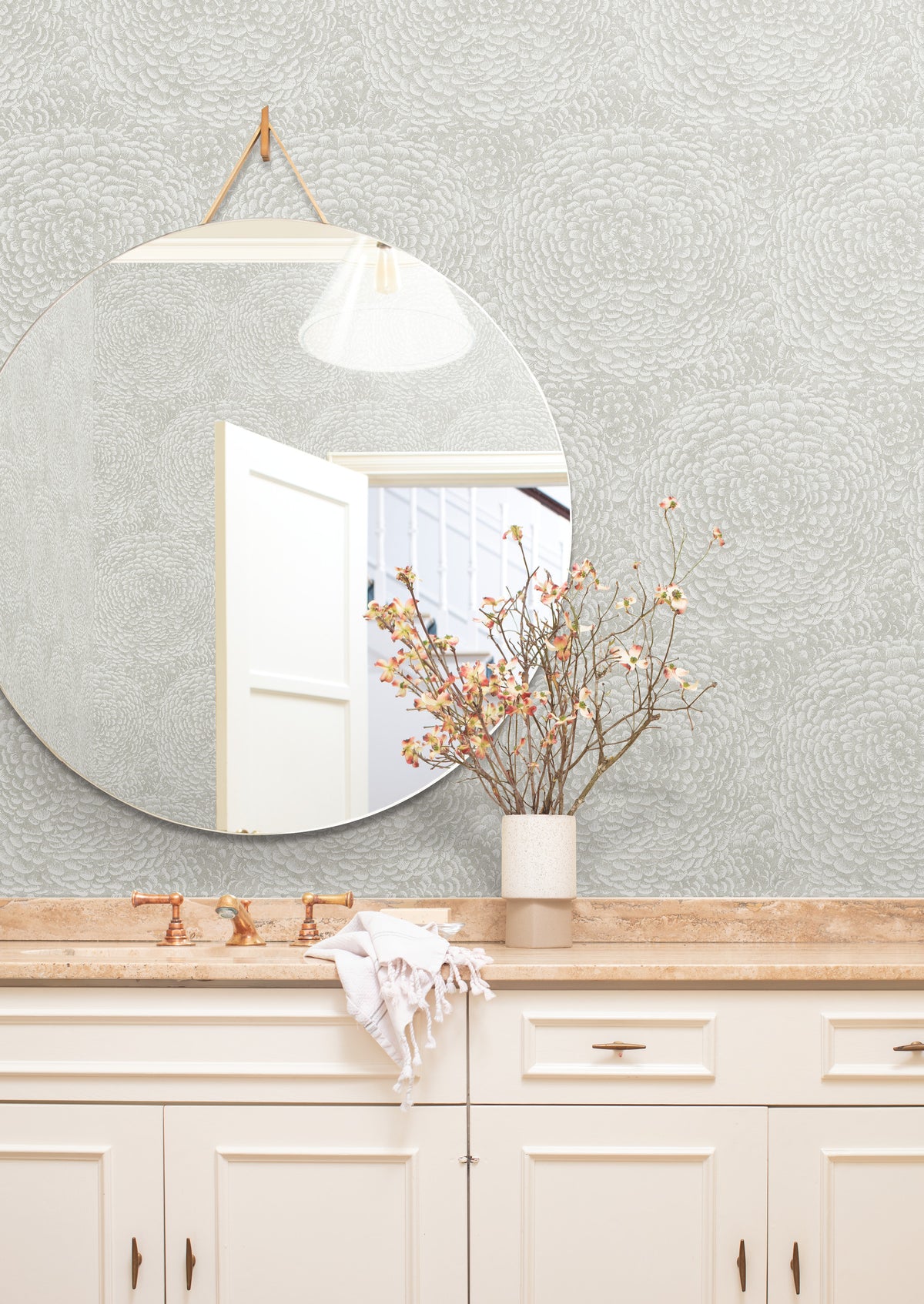 A round mirror above a beige countertop reflects a white door. A vase with branches and flowers sits on the counter, next to a white towel draped over the sink. The wall is adorned with Floret Wallpaper by York Wallcoverings, adding a subtle, textured pattern to the space.