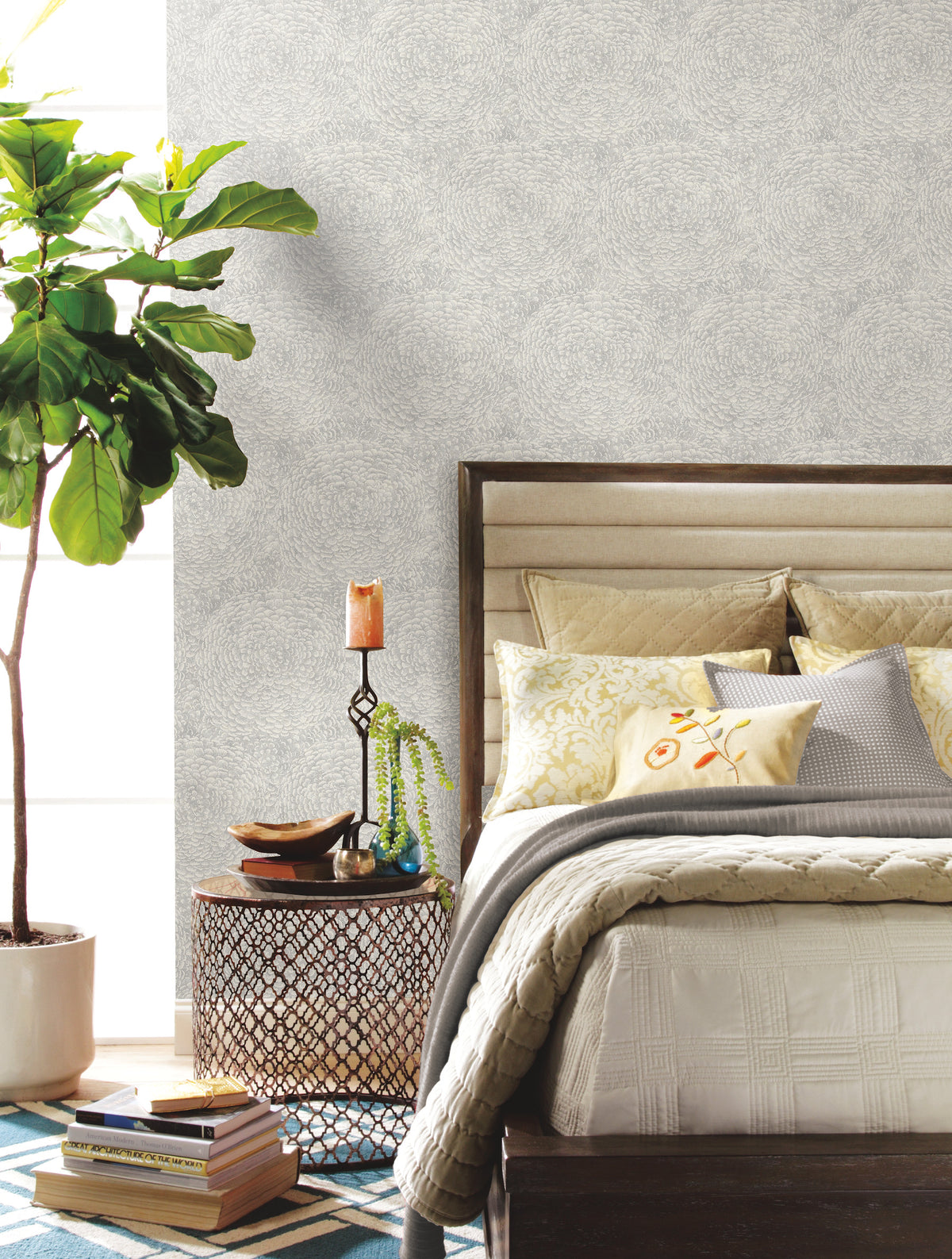 A cozy bedroom featuring York Wallcoverings’ Floret Wallpaper, a bed adorned with decorative pillows, a side table displaying a candle, plant, and books, and a large potted plant by the window. The space showcases a neutral color palette complemented by a geometric rug.